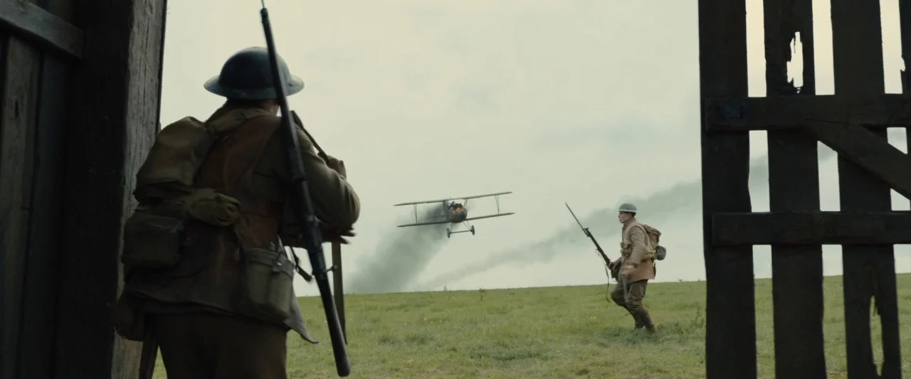 1917 movie still: wide — An open grassy field under a grey, overcast sky framed by a wooden structure. Two soldiers in period uniforms watch a…