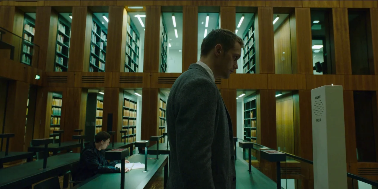 Mute (2018) movie still: medium — A modern library interior features tall wooden paneling and integrated shelf alcoves. A man stands in the…