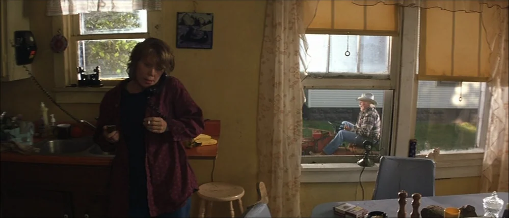 The Straight Story (1999) movie still: medium — A 1990s-style yellow kitchen features a wall-mounted telephone and a small dining table. A man in a…