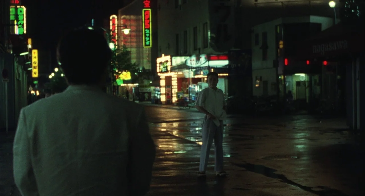 The Discarnates (1988) movie still: medium — A wet asphalt street at night is lined with illuminated Japanese business signage. A man in the…