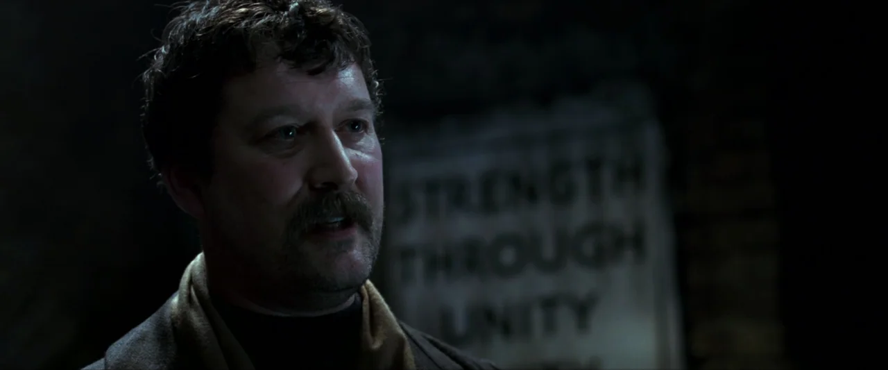 V for Vendetta (2006) movie still: closeup — A dimly lit indoor space features a wall sign displaying text. A man with a mustache talks while…