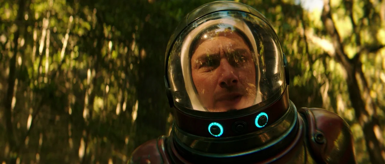 Wish I Was Here (2014) movie still: closeup — A man wearing a space helmet stands within a densely wooded area. Sunlight streaks through the canopy…