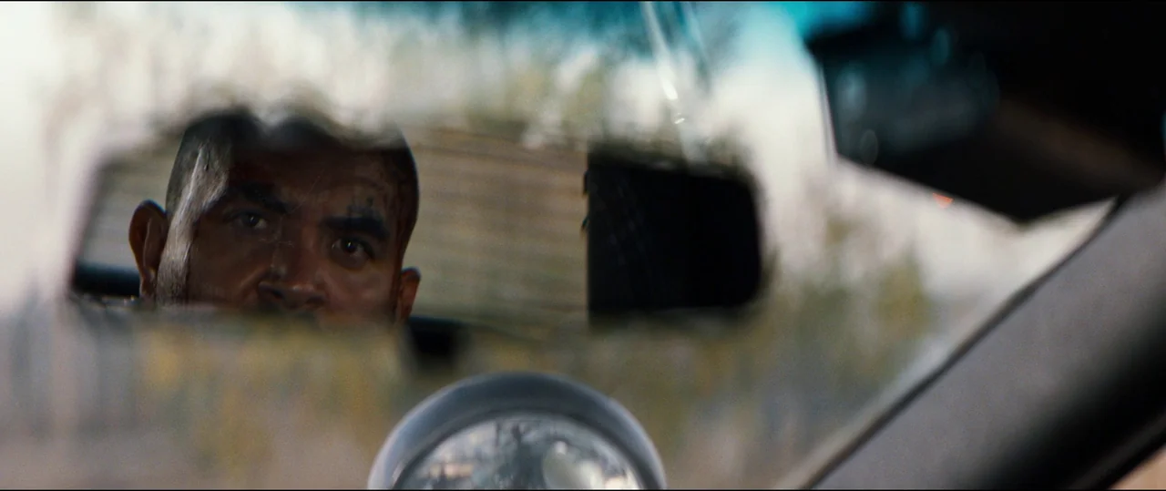 El Chicano (2018) movie still: closeup — The interior of a vehicle seen through the reflection of a rearview mirror. A man gazes forward with a…