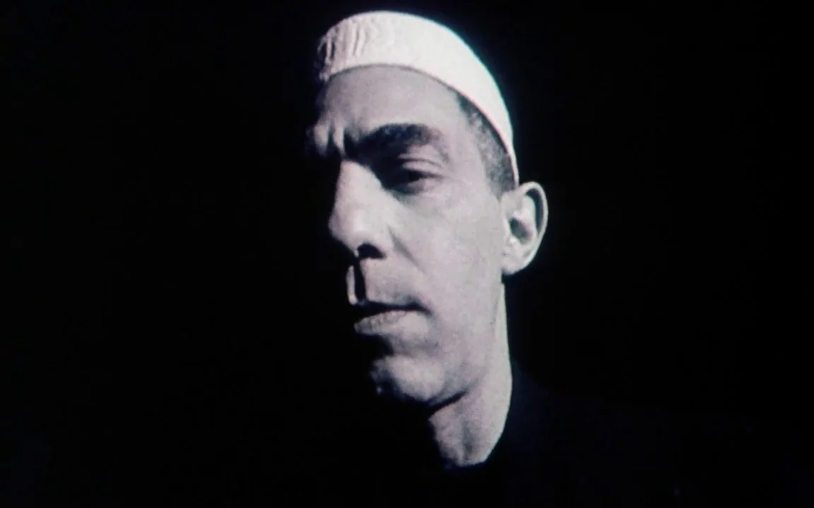 The Last of England (1987) movie still: closeup — A man stands against a pitch-black background with no visible surroundings. He wears a small white…