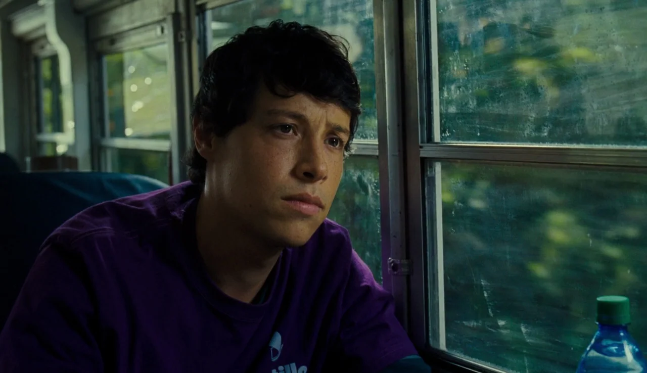 Our Brand is Crisis (2015) movie still: medium — A passenger sits by a window inside a bus with a view of greenery passing. The young man stares…