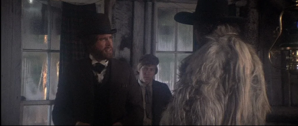 McCabe & Mrs.Miller (1971) movie still: medium — A dimly lit, rustic interior features wooden walls and frosted window panes. A bearded man in a…