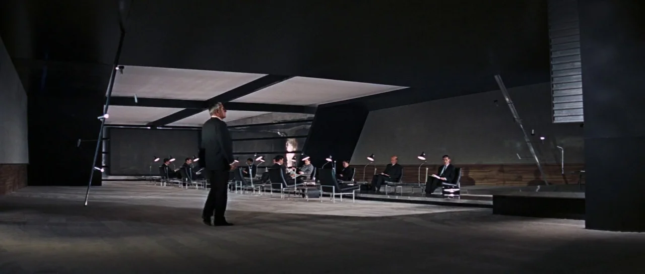 Thunderball (1965) movie still: wide — A spacious, modern hall features black walls, a flat ceiling, and rows of chairs. A man walks across the…