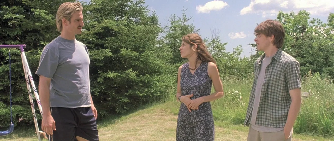 In the Bedroom (2001) movie still: fullbody — A grassy lawn features three people standing and talking near a metal swing set. Tall coniferous trees…