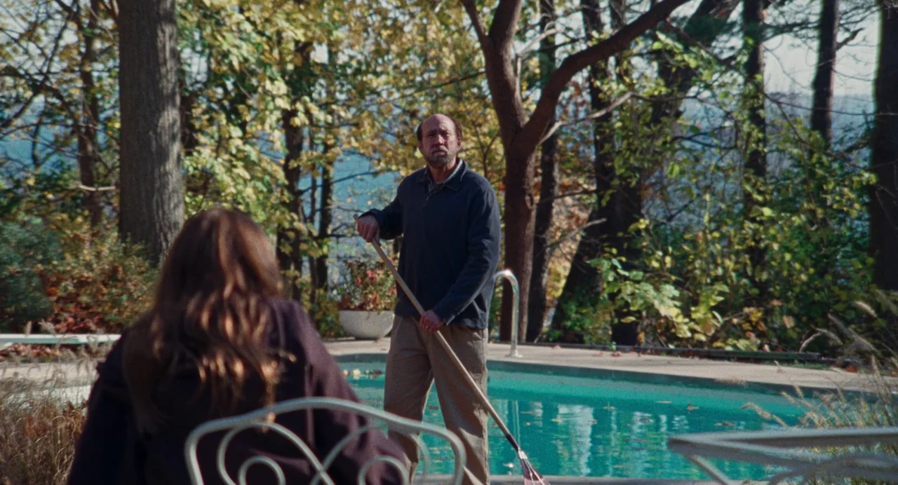 Dream Scenario (2023) movie still: medium — A patio area overlooks a backyard swimming pool surrounded by mature deciduous trees. A middle-aged man…