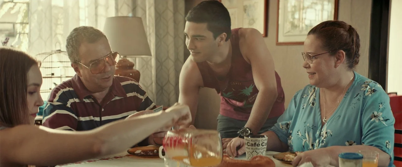 The Boy Who Killed My Parents (2021) movie still: medium — A sunlit dining room features patterned wallpaper and framed artwork on the walls. A…