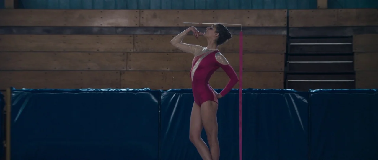 Alps (2011) movie still: fullbody — An empty gymnasium features wooden walls and blue protective mats along the base. A young gymnast stands in…