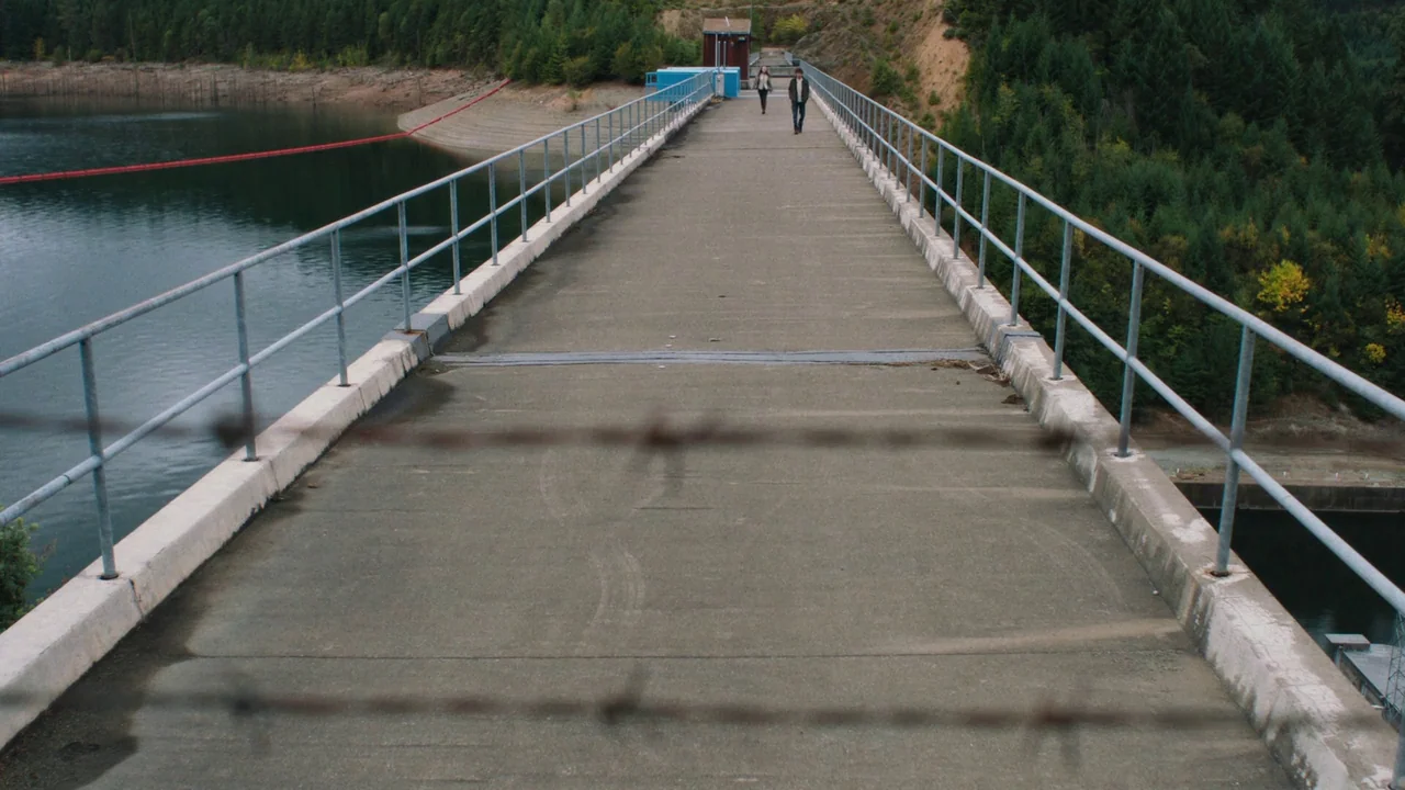 Night Moves (2013) movie still: wide — A concrete dam walkway stretches forward, flanked by water and dense coniferous forest. A young man and woman…
