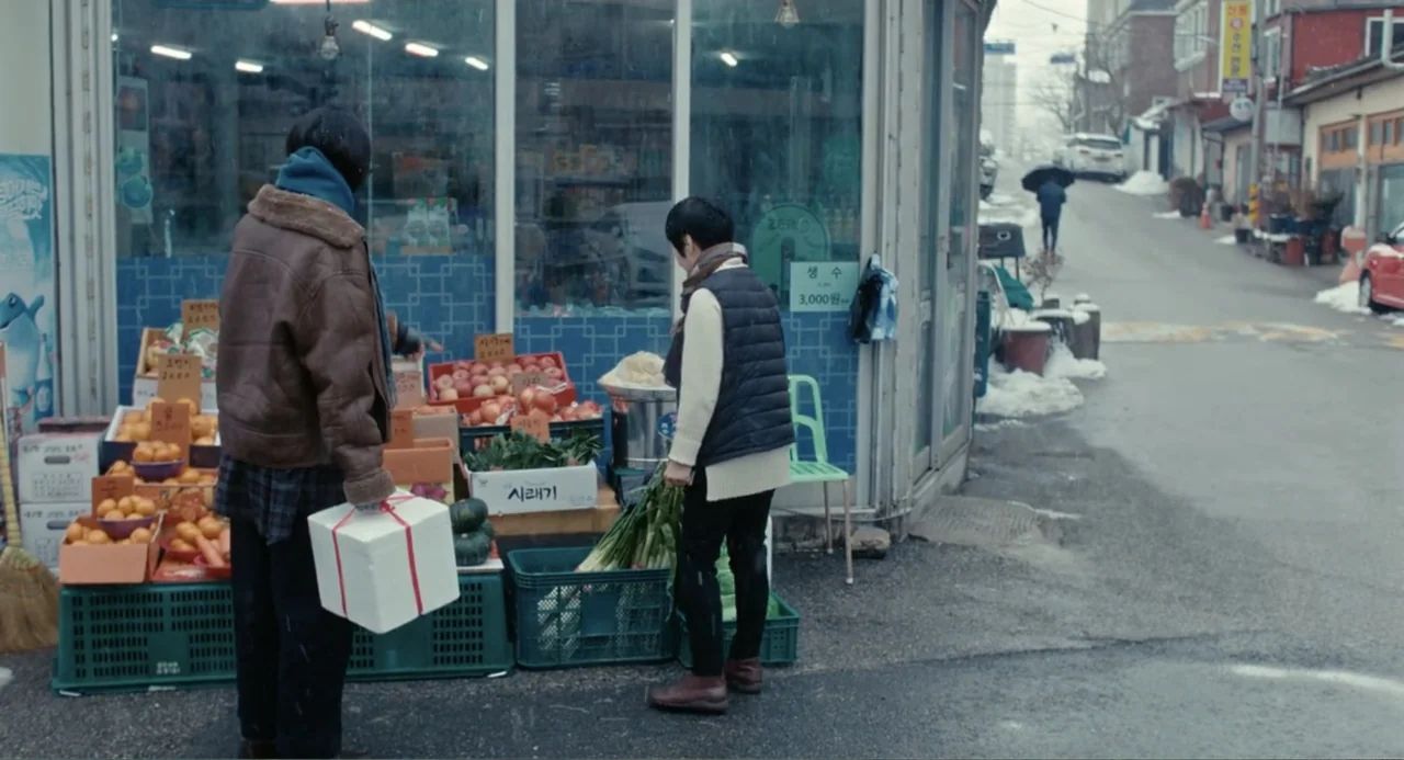 Winter in Sokcho (2025) movie still: fullbody — A corner convenience store features fresh produce displayed in plastic crates on the sidewalk. Two…