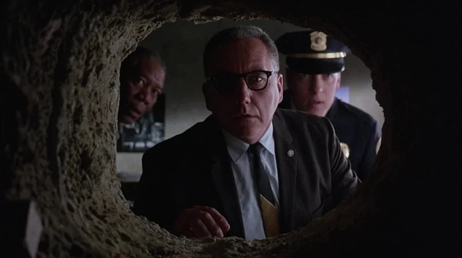 The Shawshank Redemption (1994) movie still: medium — Three men stand inside a dimly lit room behind a wall with a hole. They look through the…