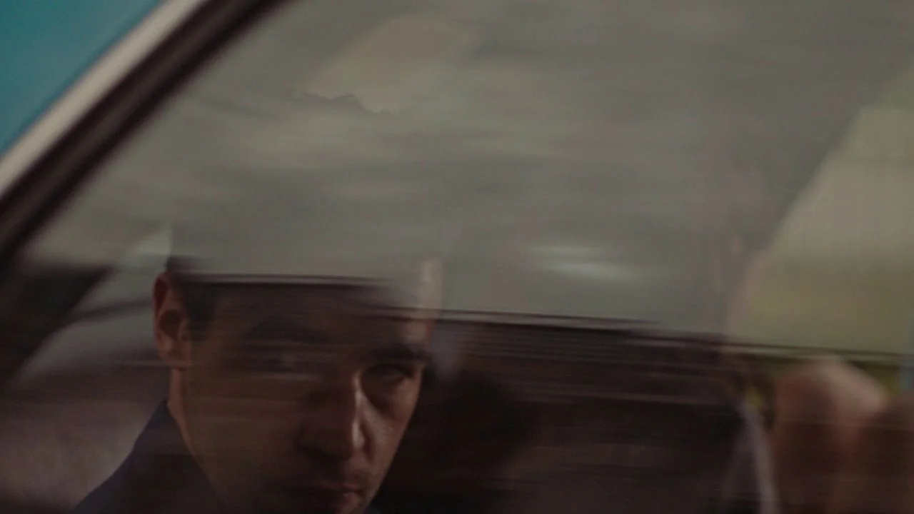 Possessor (2020) movie still: closeup — A man sits inside a vehicle with motion blur visible through the glass. The reflection on the car window…
