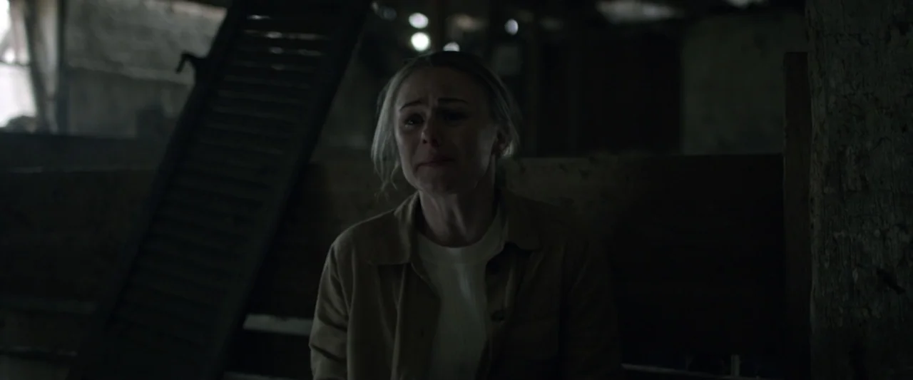 Motherly (2022) movie still: medium — A dark, weathered barn interior with wooden structural beams and muted textures in the shadows. A woman with…
