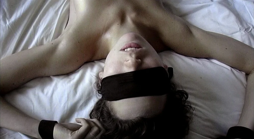 9 Songs (2004) movie still: closeup — A person lies on white bedsheets with their arms spread outward. A black blindfold covers the eyes and black…