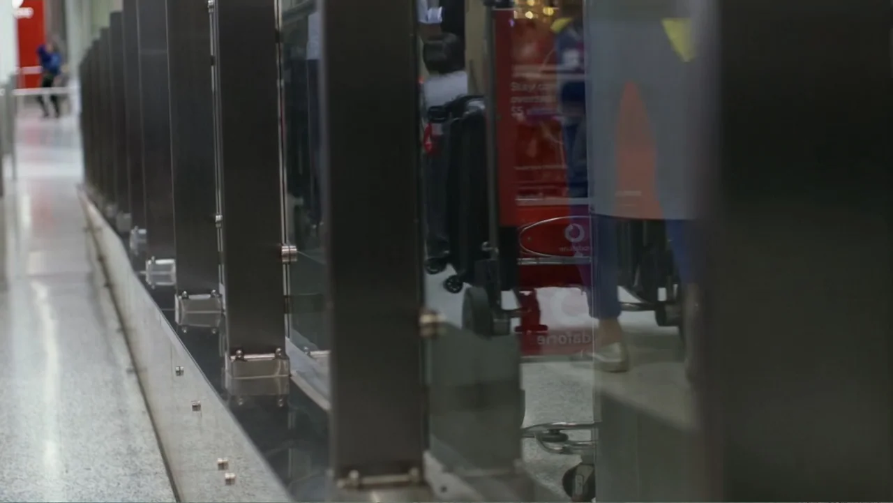 Escape From Saudi (2019) movie still: medium — A polished glass partition stands in a brightly lit indoor transit area. Luggage carts are partially…