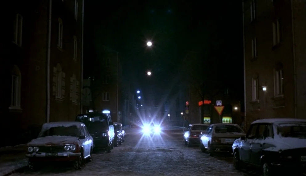 Night on Earth (1991) movie still: wide — A quiet snowy city street at night is framed by two rows of multi-story buildings. An oncoming car with…