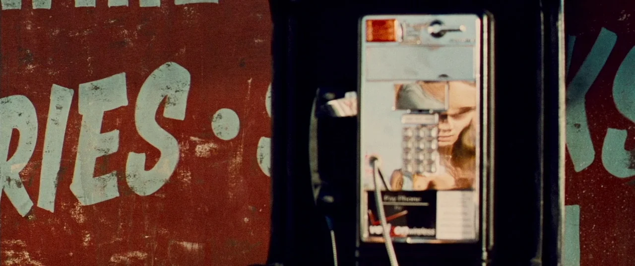 Carriers (2009) movie still: closeup — A public payphone is mounted against a dark red wall with painted blue-green lettering. The device captures a…