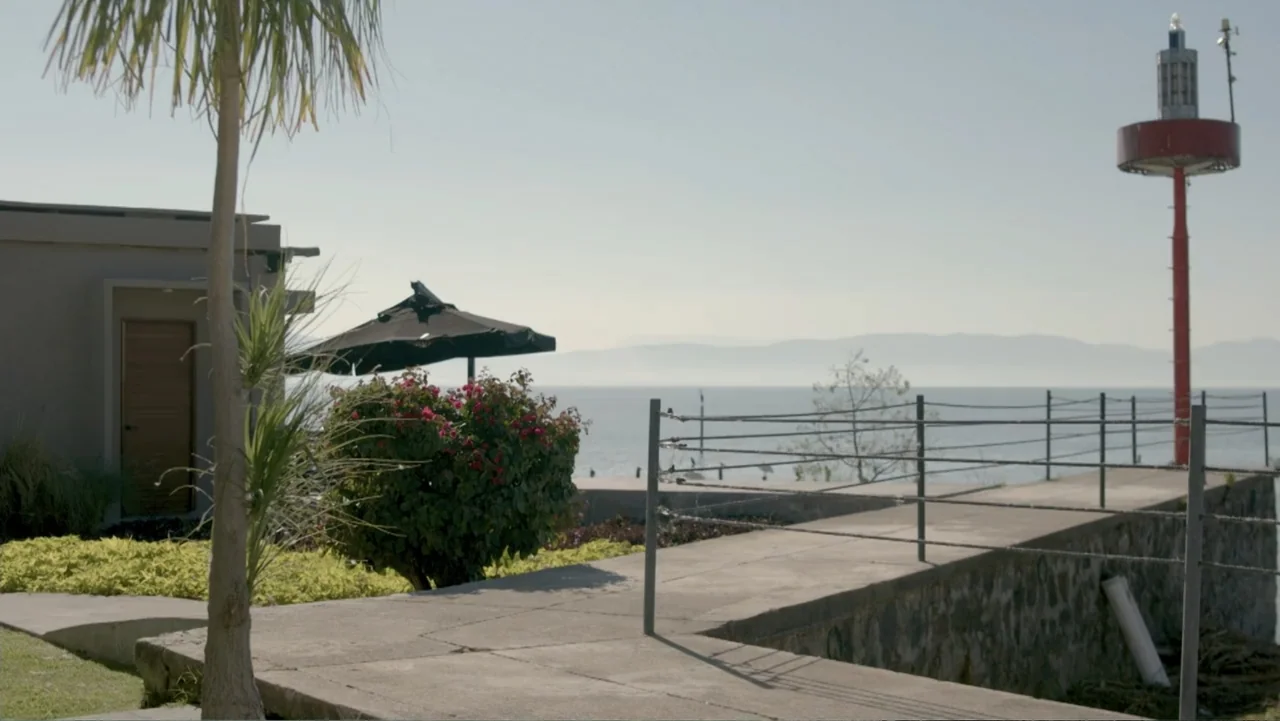 Por Amor al Money (2023) movie still: establishing — A seaside pier with a tall red beacon, palm tree, and black patio umbrella. The structure…