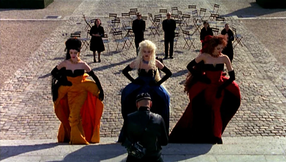 The Tango Lesson (1997) movie still: fullbody — A stone paved courtyard contains rows of metal cafe chairs and standing figures. Three women wearing…