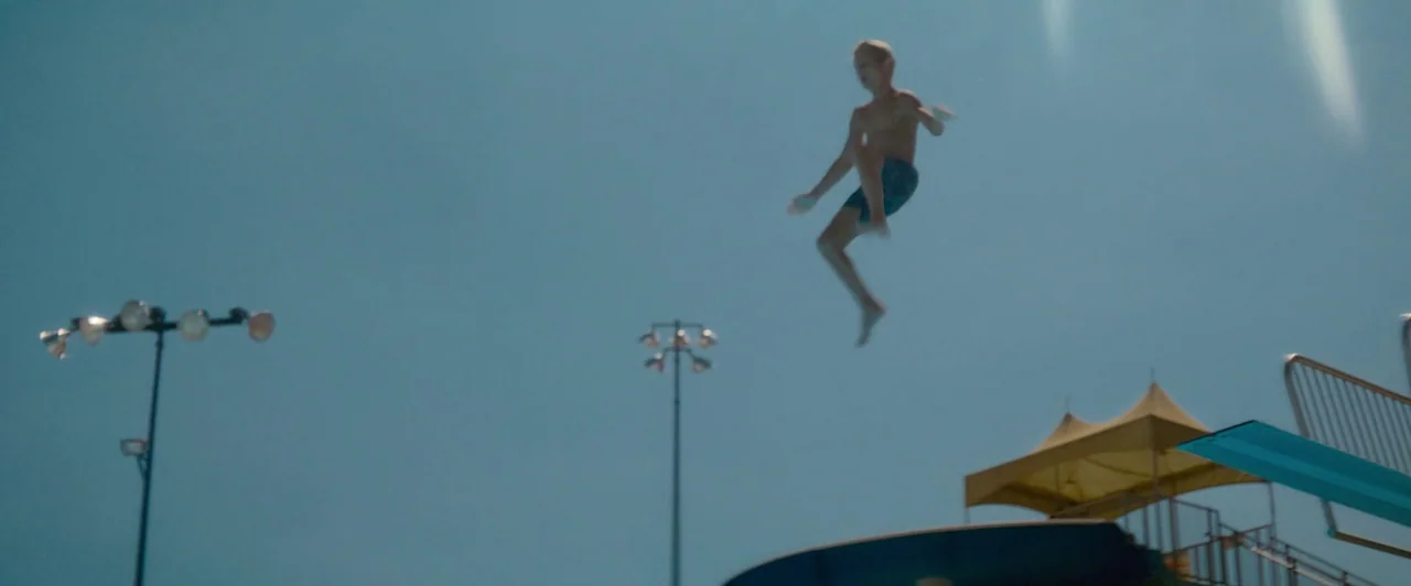 Snack Shack (2024) movie still: wide — An outdoor aquatic facility featuring a metal diving board and a yellow canopy tent. A young boy performs a…