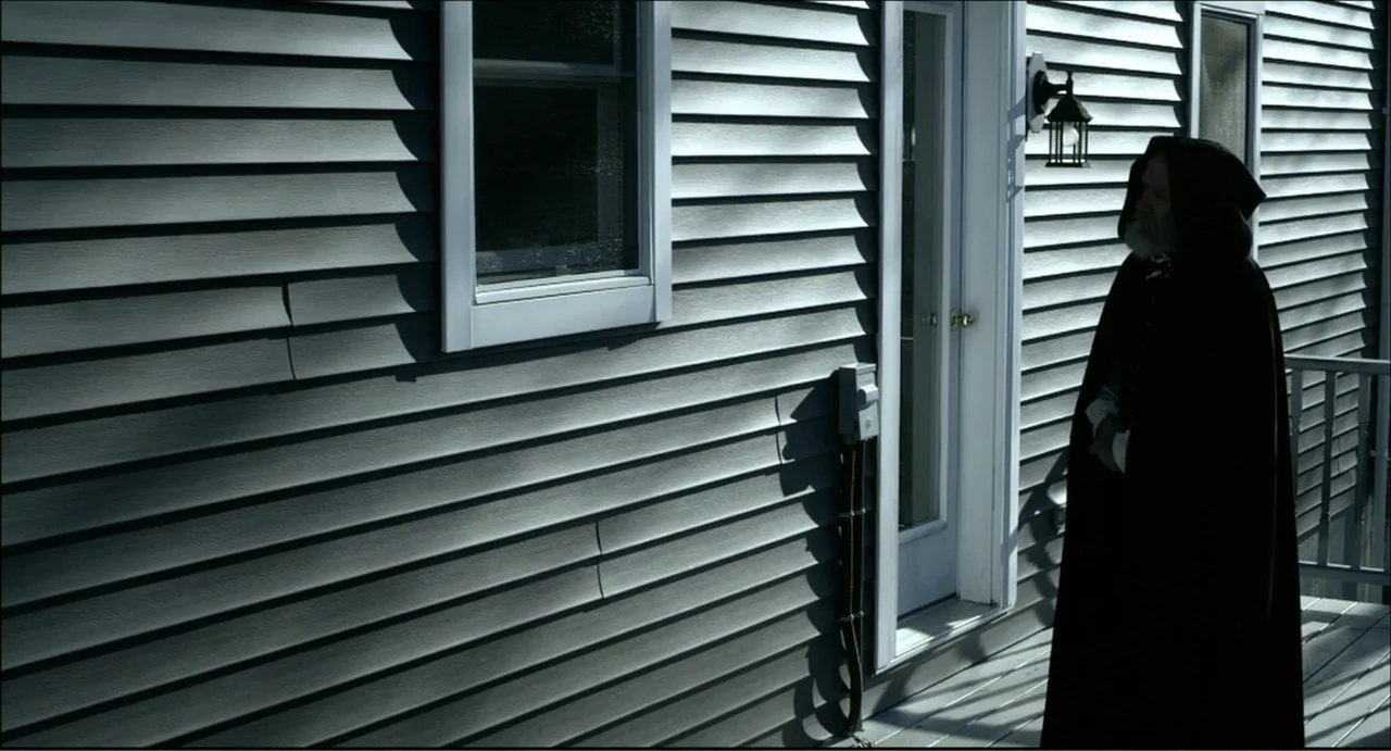The Secret Village (2013) movie still: fullbody — A man in a hooded cloak stands on a porch with vinyl siding. The man stands still against the wall…