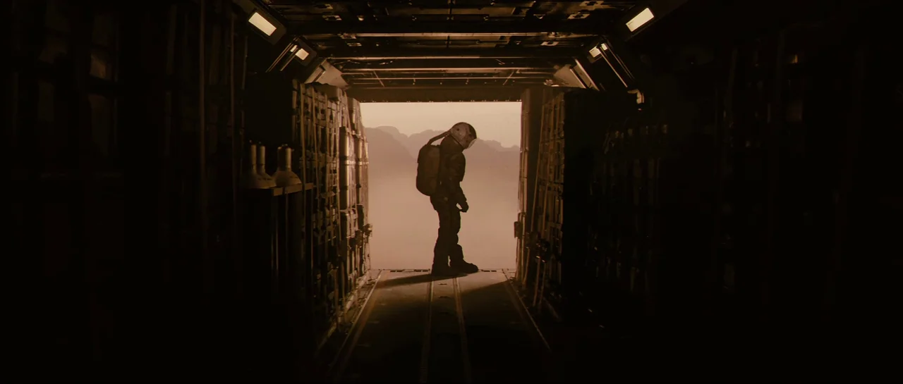 The Last Days On Mars (2013) movie still: fullbody — A dark, industrial cargo hold is lined with stacks of rectangular storage containers. An…