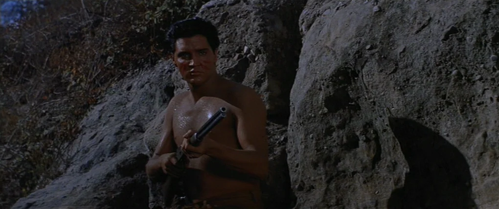 Flaming Star (1960) movie still: medium — A rugged outdoor setting features a large rock formation under harsh sunlight. A shirtless man stands…