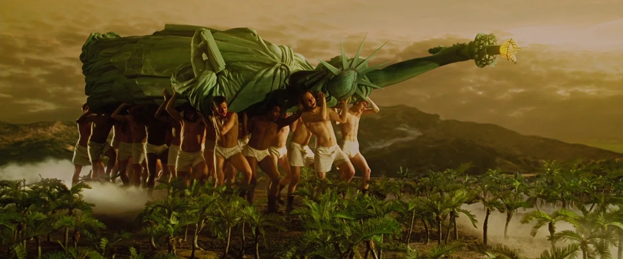 Across the Universe (2007) movie still: wide — A vast field of small palm trees sits beneath a golden sunset sky. A group of shirtless men in white…