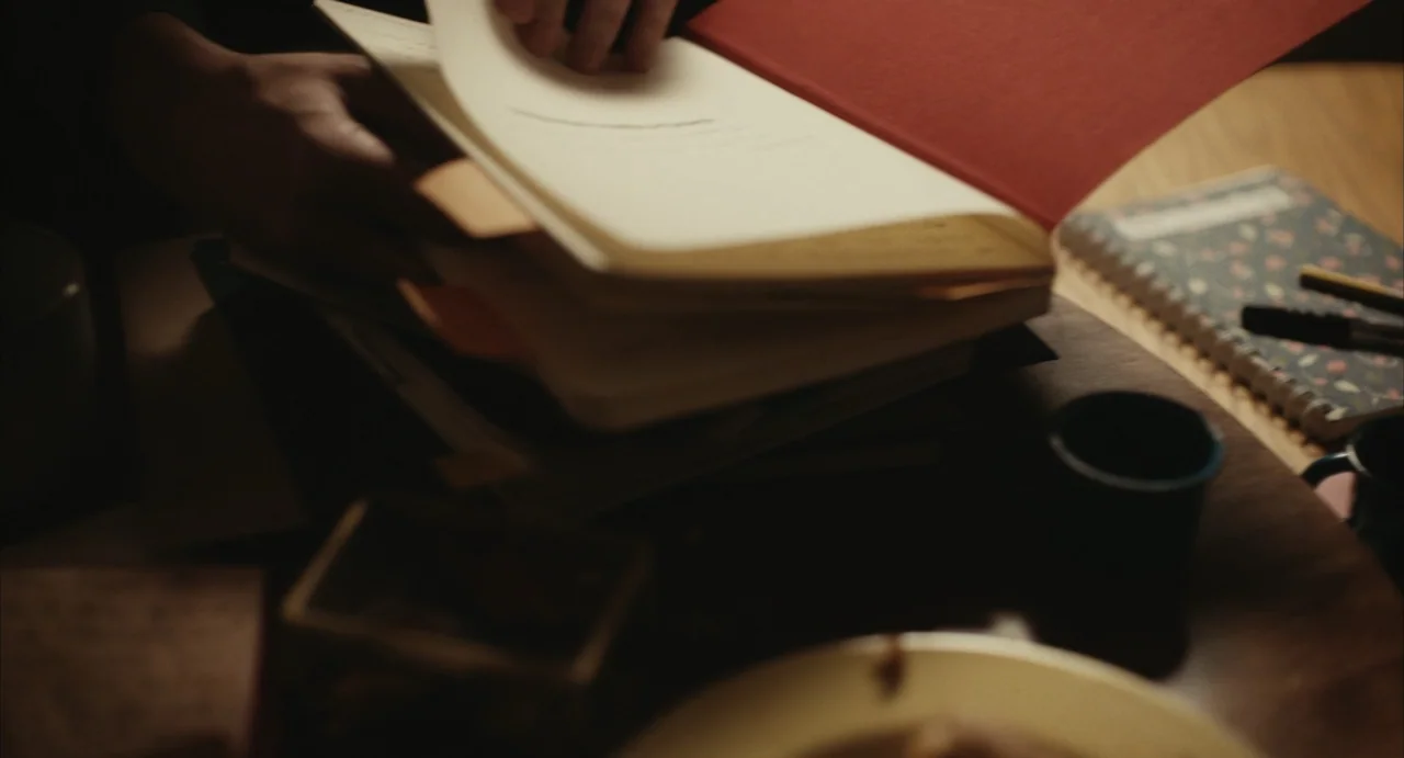 The Diary (1999) movie still: closeup — A wooden desk covered in scattered papers and a red-covered book. Hands flip through the pages of the open…