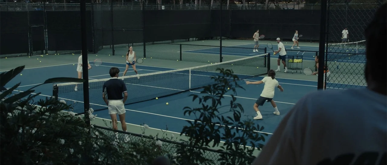 King Richard (2021) movie still: wide — Multiple tennis courts with chain-link fencing are visible during a bright daylight match. Various players…