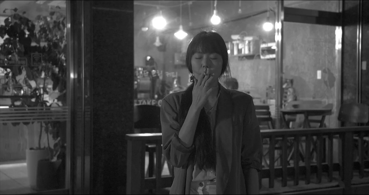 Grass (2018) movie still: medium — A woman stands on a sidewalk at night in front of a glass storefront. She holds a lit cigarette to her lips with…