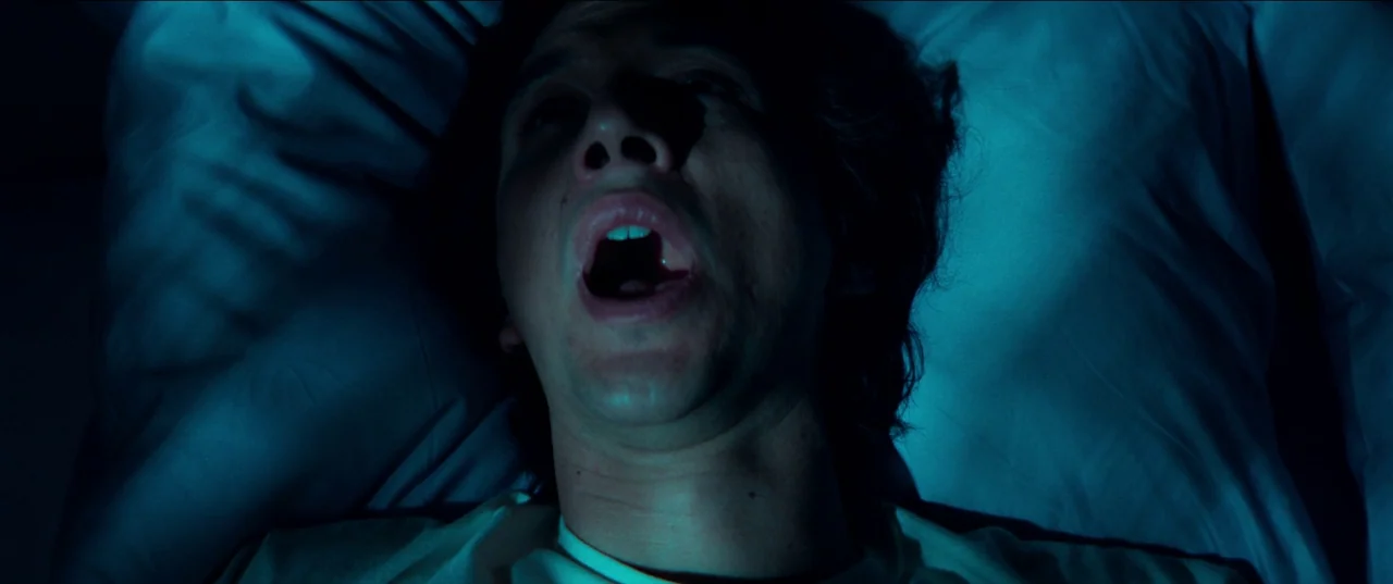 White Noise (2022) movie still: closeup — A man lies on a white pillow at night. He is illuminated by the blue light of a screen.