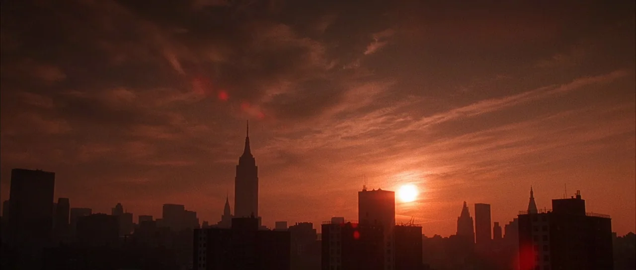 Die Hard with a Vengeance (1995) movie still: establishing — A dense urban skyline is silhouetted against a deep red sunset. The sun sits low behind…