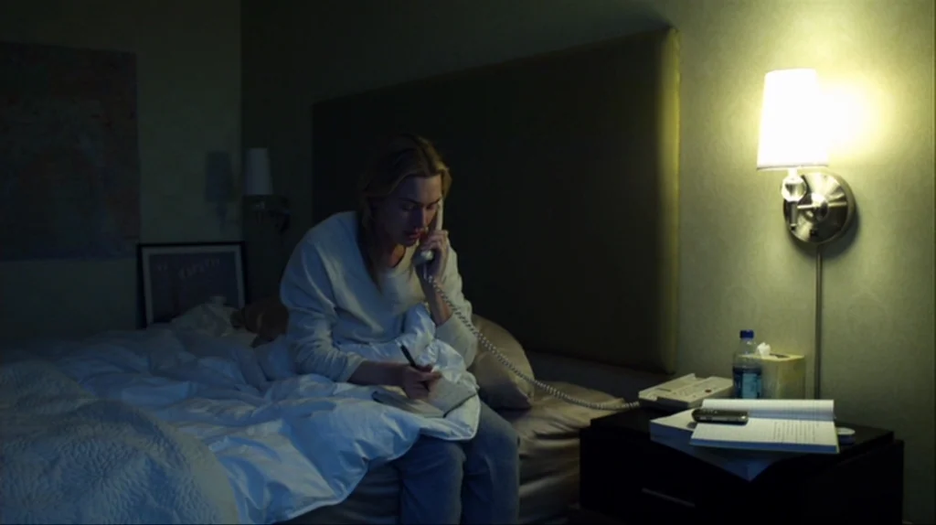 Contagion (2011) movie still: medium — A dimly lit room contains a bed with a large upholstered headboard and a nightstand. A woman sits on the edge…