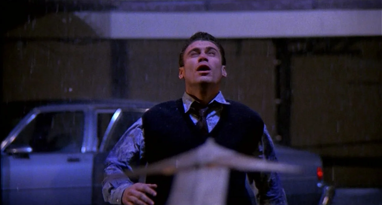 Raising Cain movie still: medium — A man stands outdoors in the rain at night in front of a parked car. He looks upward with a wide-eyed, distressed…