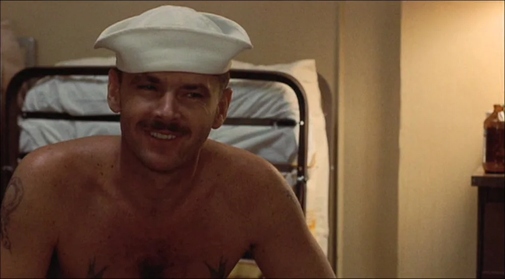 The Last Detail (1973) movie still: medium — A spartan hospital room features a metal bed frame against a plain beige wall. A bare-chested man with…
