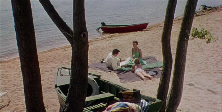 Magnificent Obsession (1954) movie still: wide — A sandy beach shoreline features three people relaxing on a blanket near the water. Two small…