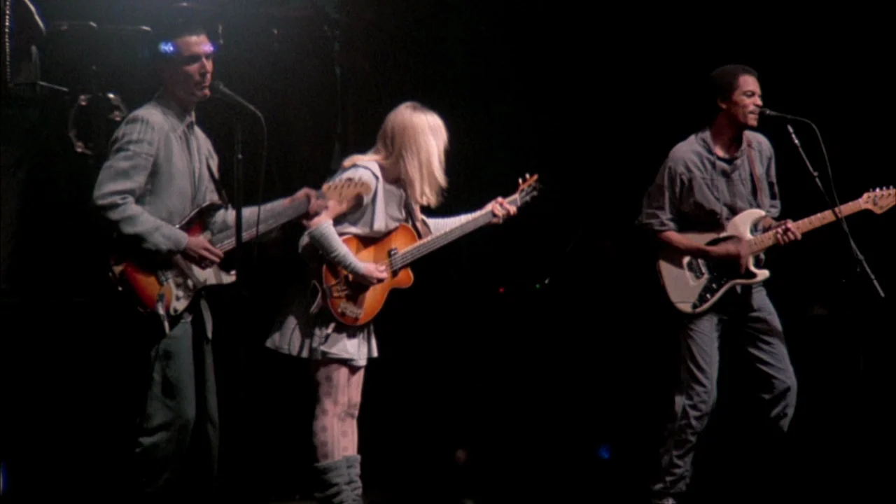 Stop Making Sense (1984) movie still: medium — A dark concert stage hosts three musicians performing under bright directed spotlights. They play…