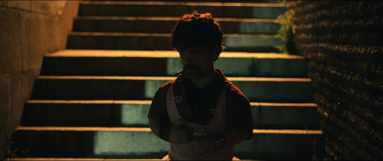 The Toxic Avenger (2023) movie still: medium — An outdoor staircase with concrete steps and stone walls lit at night. A man with a mustache stands…