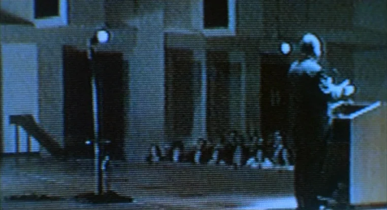 Schizopolis movie still: wide — An auditorium with a sparse audience in the dark background behind a speaker. A man stands at a podium in the…