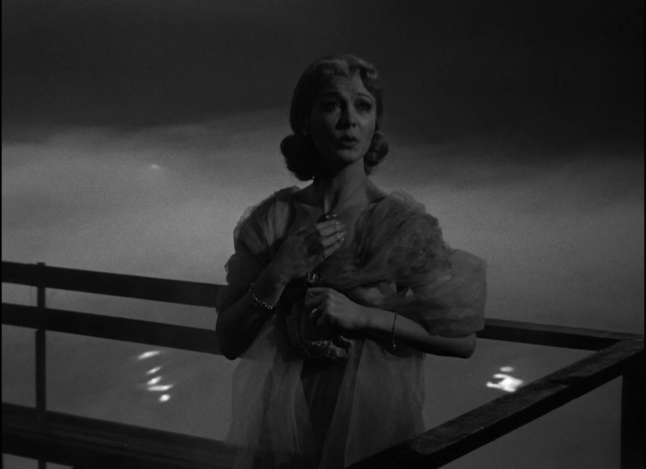 A Streetcar Named Desire (1951) movie still: medium — A woman stands on a balcony at night overlooking a dark body of water. She grips a clutch…