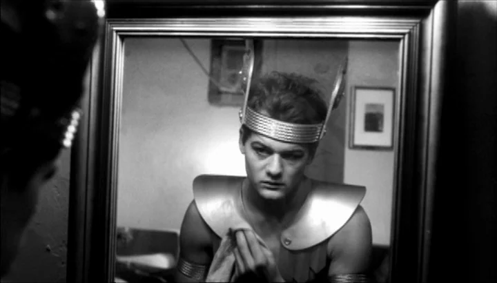 The American Astronaut (2001) movie still: closeup — A man in theatrical costume stares at his reflection in a framed wall mirror. The setting is a…