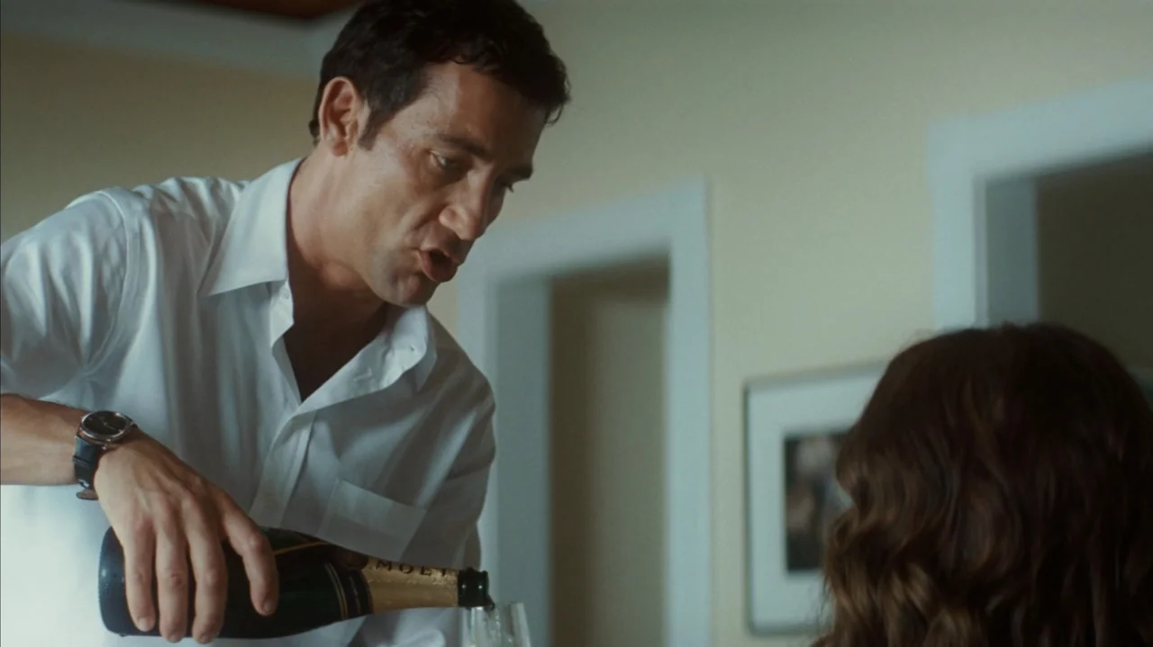 Duplicity (2009) movie still: medium — A light-toned interior room contains a man and woman at a table together. A man in a white shirt pours…