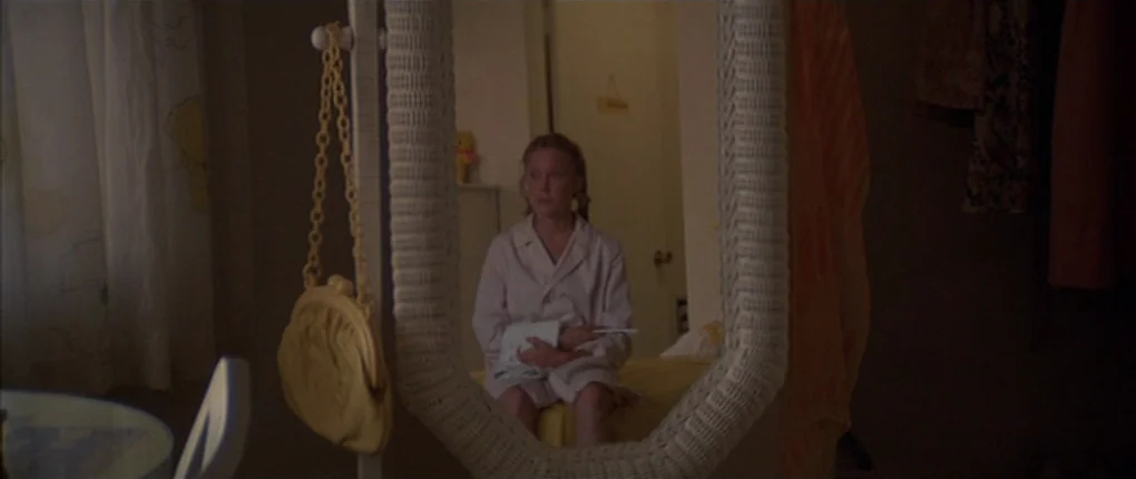 3 Women (1977) movie still: medium — A dimly lit room reflected in a white woven-frame mirror displays a young woman. She sits quietly on a yellow…