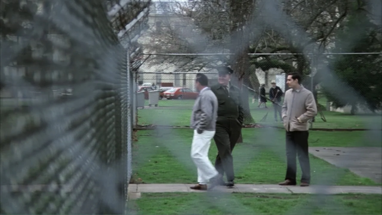 One Flew Over the Cuckoo's Nest (1975) movie still: wide — A fenced-in outdoor area with a paved path across a green grass lawn. Three men walk…