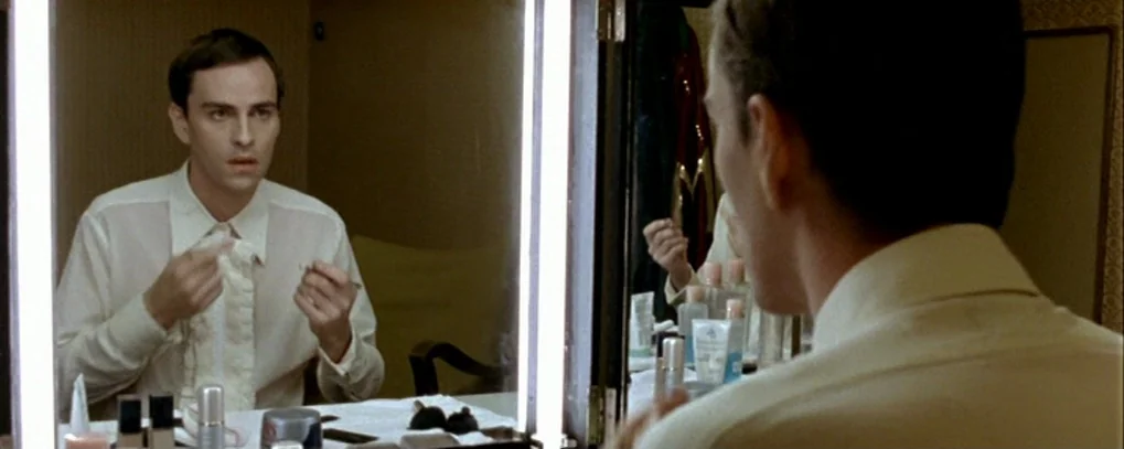 Calvaire (2004) movie still: medium — A man looks into a vanity mirror inside a brightly lit dressing room. The man holds small objects near his…