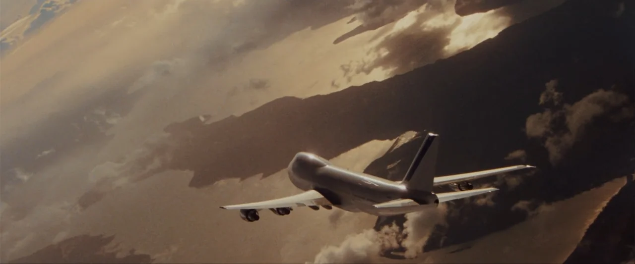 Unfinished Business (2015) movie still: wide — A passenger airplane is seen from the rear flying over a coastline at altitude. The plane travels…